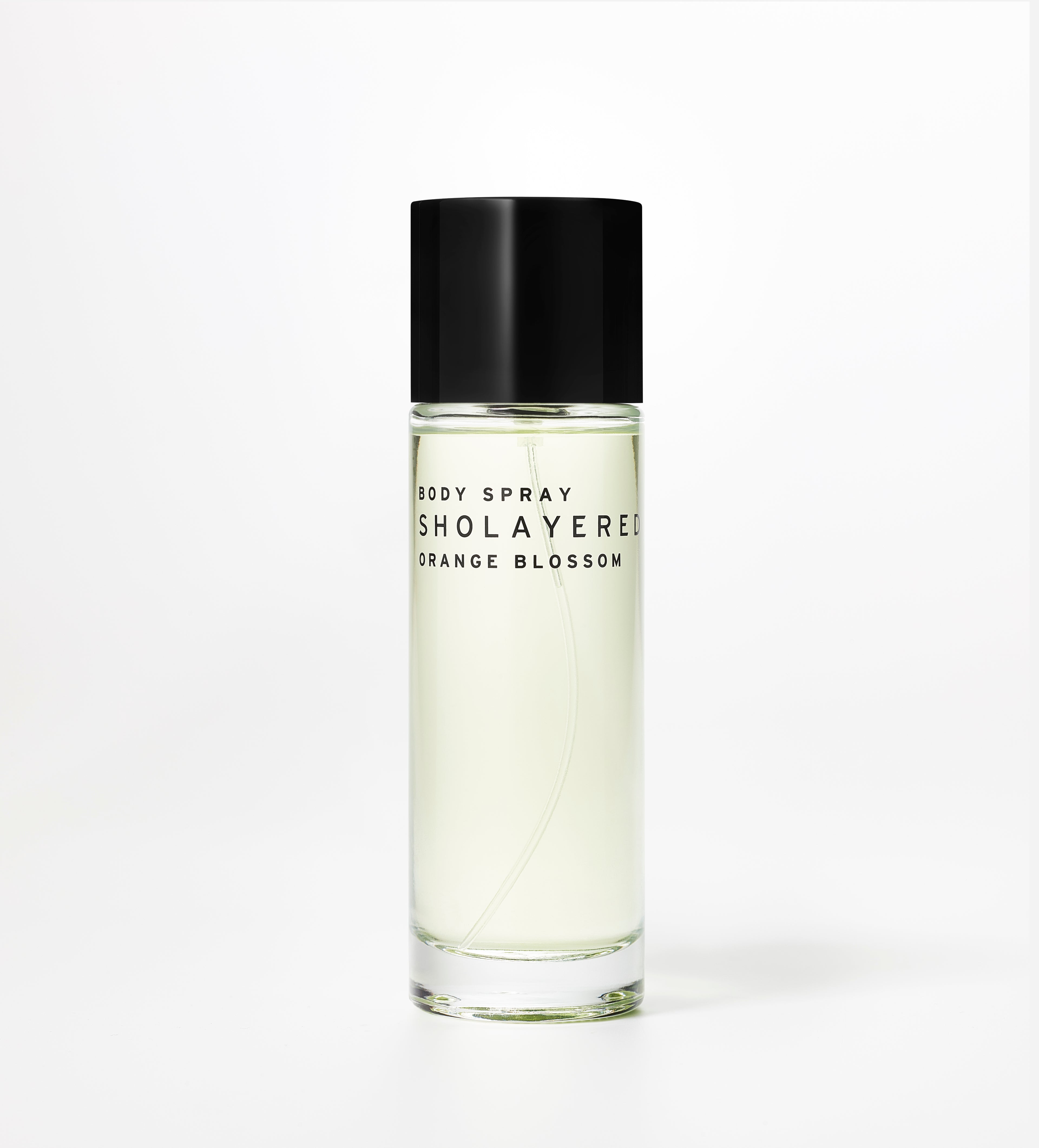 A clear glass bottle of SHOLAYERED Fragrance Body Spray with a black spray nozzle, labeled with 'Orange Blossom'. The liquid appears to be clear or yellowish.